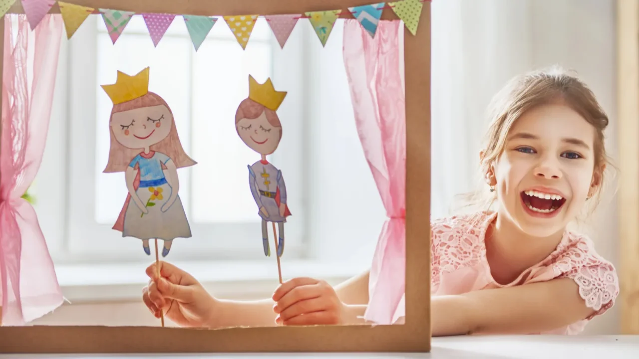 A girl playing with a DIY puppet theater made up of cardboard, fabric curtain, and paper puppets.