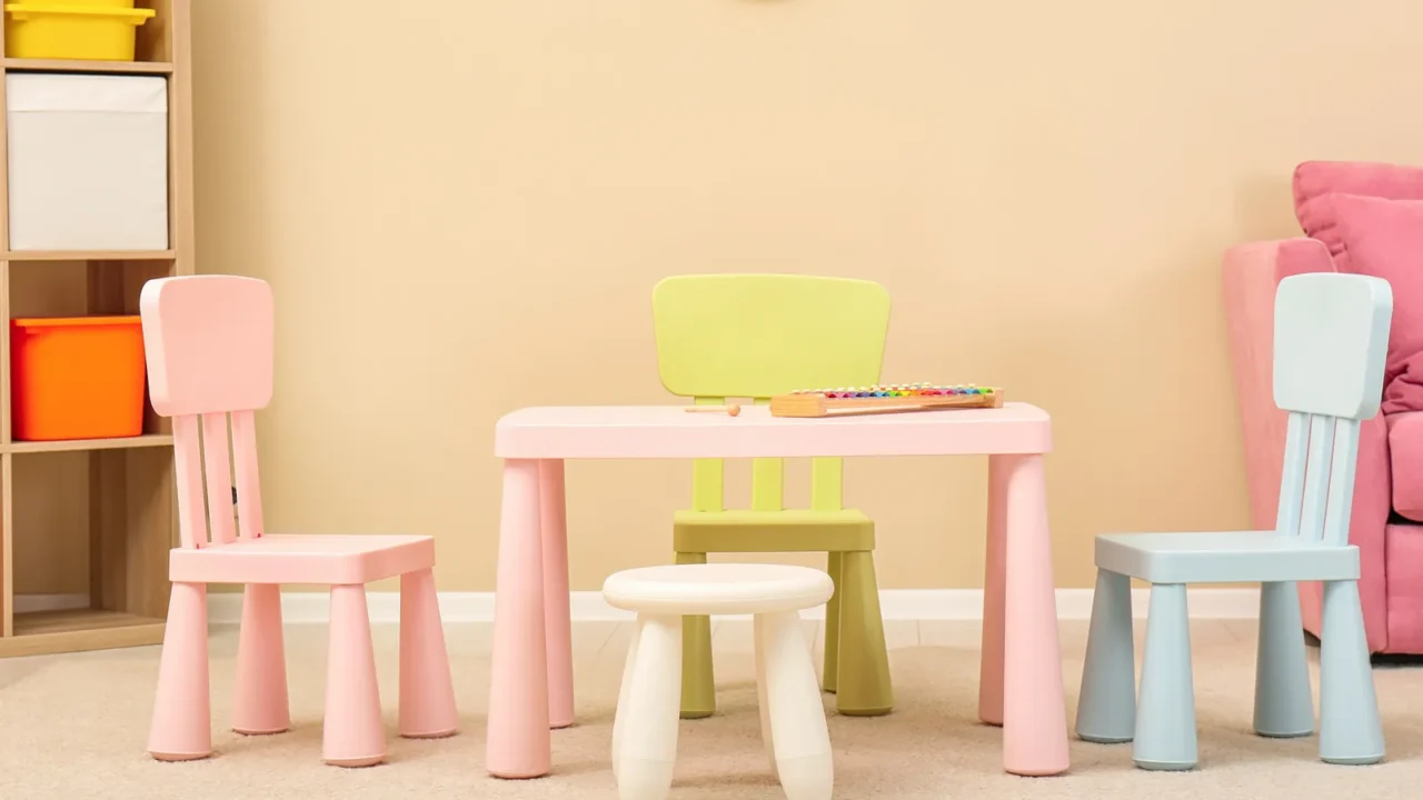 A room with pink table and pink, blue, green chairs, and a wooden shelf beside a peach wall.