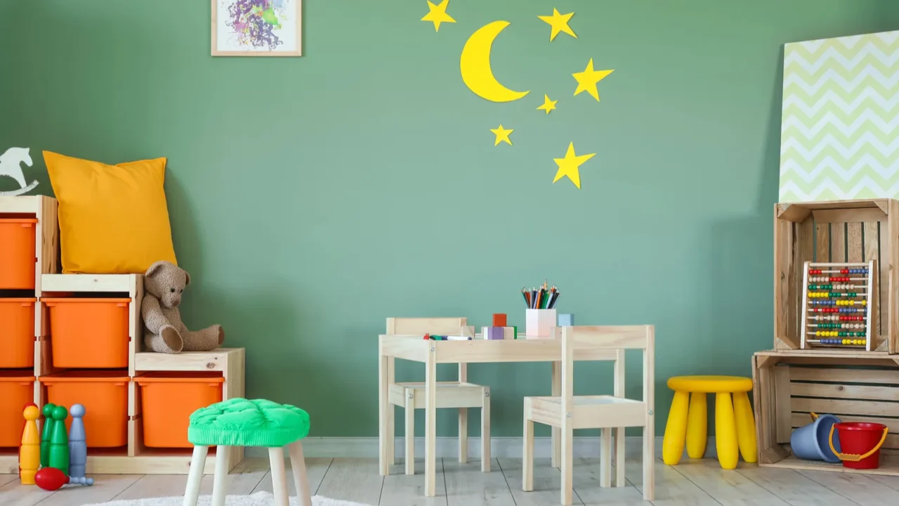 A playroom interior with wooden crates shelf, wooden table and chair, toys, and moon and stars pasted on a sage green wall.