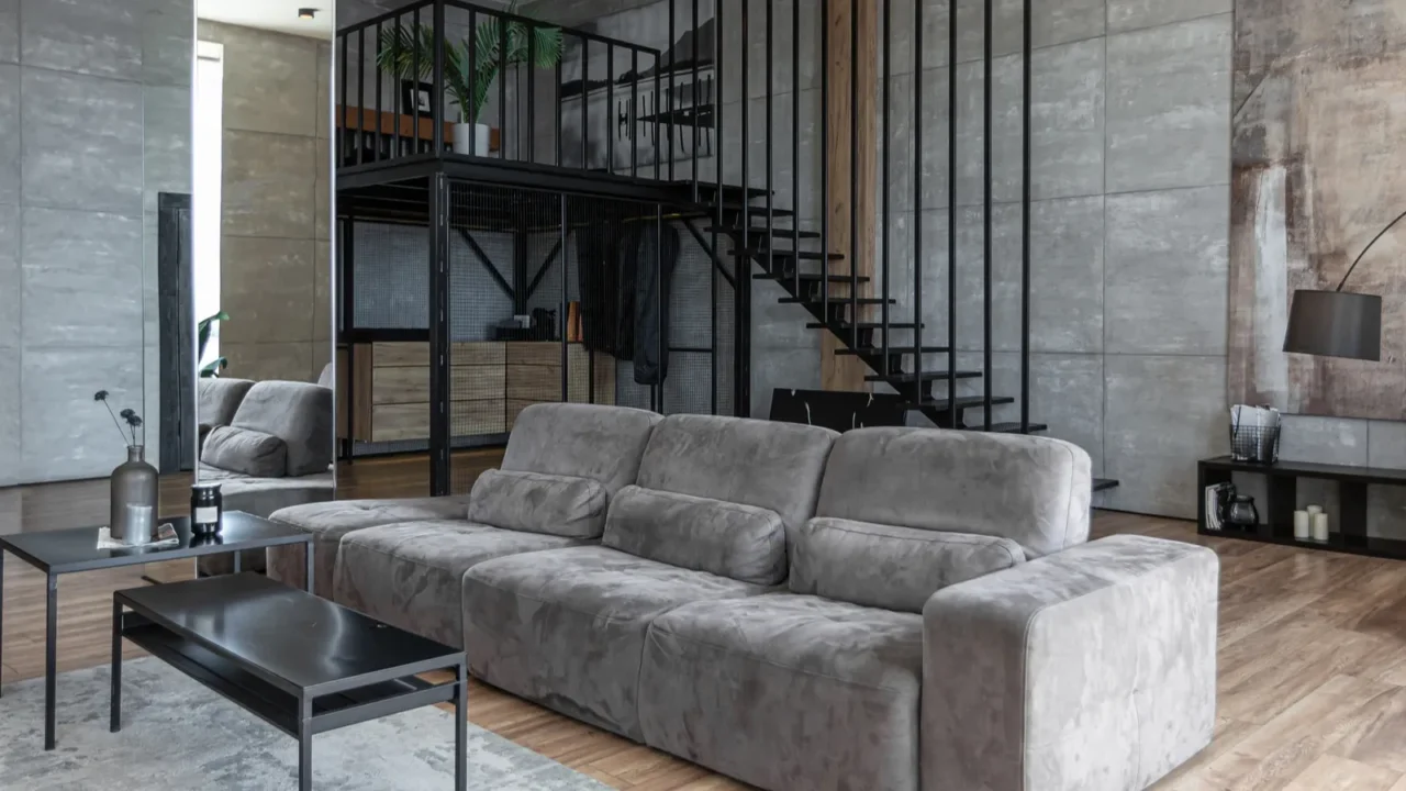 A sleek gray living room with a plush sofa, a glass coffee table, and a staircase in the background.