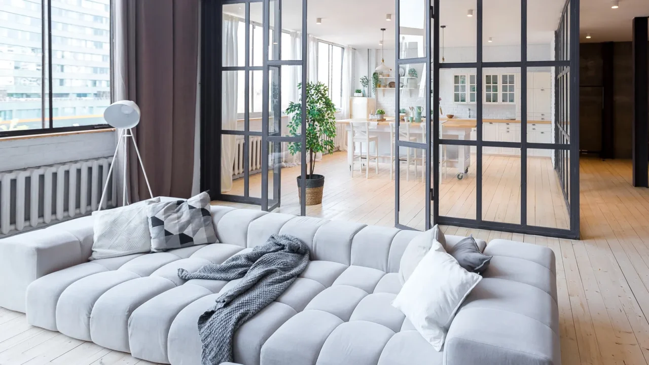 A modern living room with a gray sofa, large windows, and a view of a kitchen area.