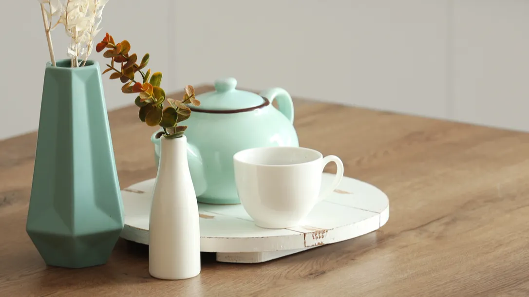 A mint green teapot and a white cup on a wooden surface with decorative vases.