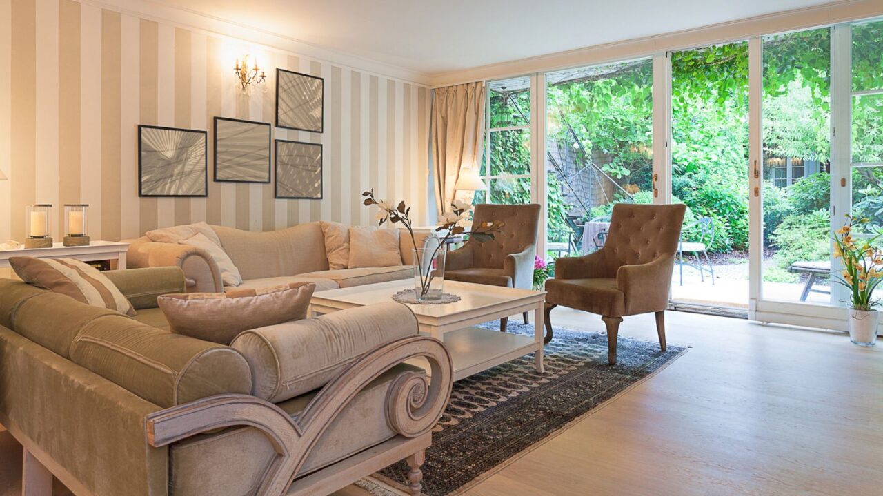 A cozy living room features a beige sofa, decorative pillows, and large glass doors leading to a green garden.