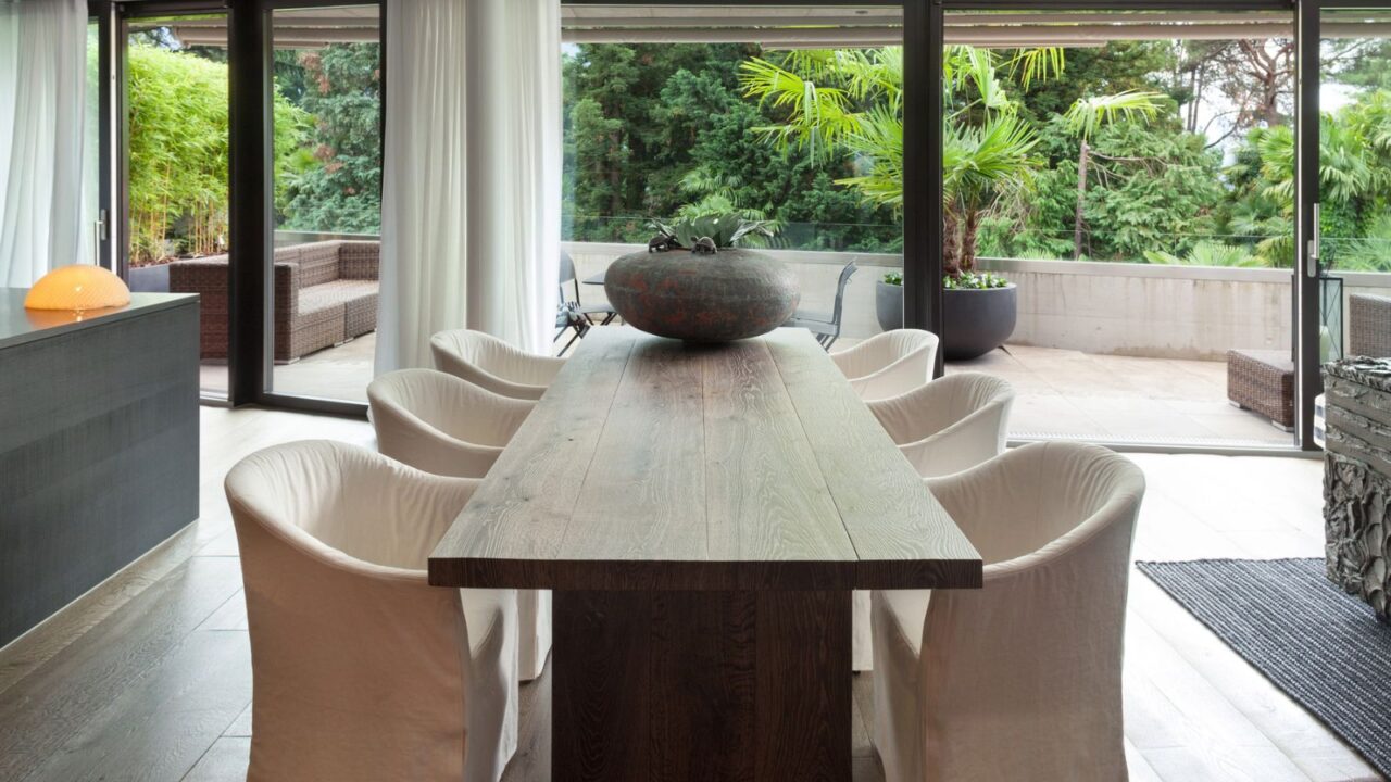A modern dining area showcases a long wooden table with comfortable chairs and large windows overlooking greenery.