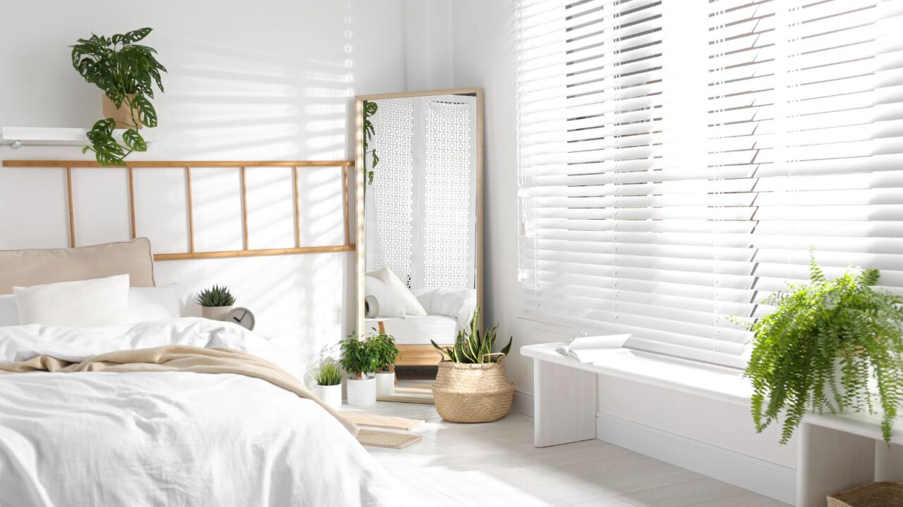 A neutral bedroom interior with white bedding, houseplants, and a full-length mirror placed in a corner near the window.