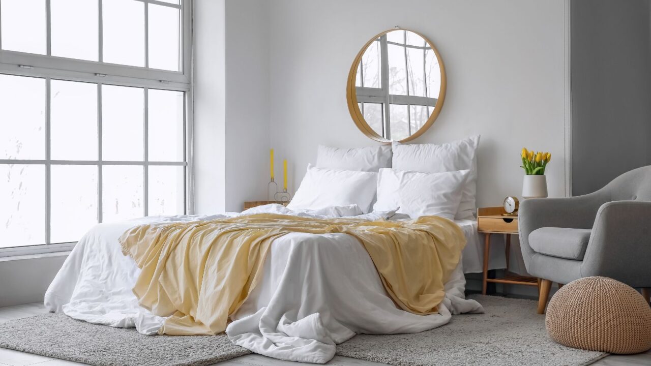 A bedroom interior with a cozy bed, gray sofa, pouf, rug, and a mirror on the wall reflecting natural light coming through the windows.