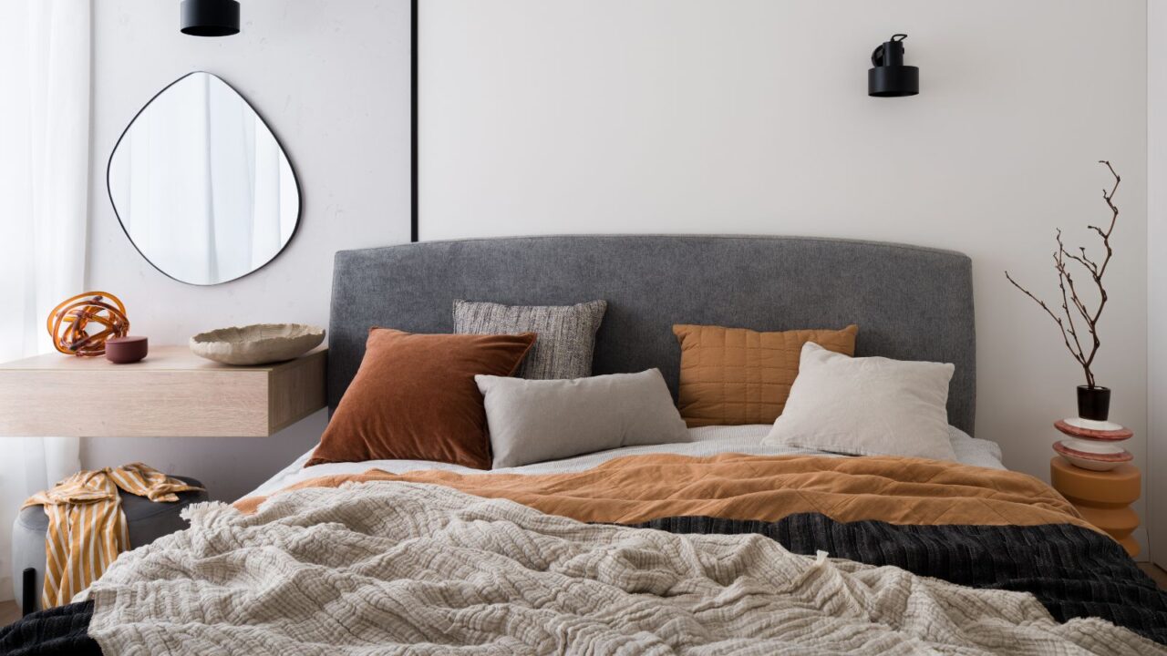 A stylish bedroom interior with a cozy bed, wooden floating nightstand, vase with dried branches, and a unique mirror on the wall.