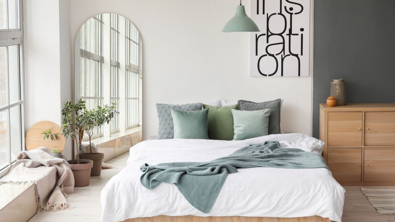 A bedroom interior with cozy bed with comfortable bedding, full standing mirror, artwork, and houseplant near a window.