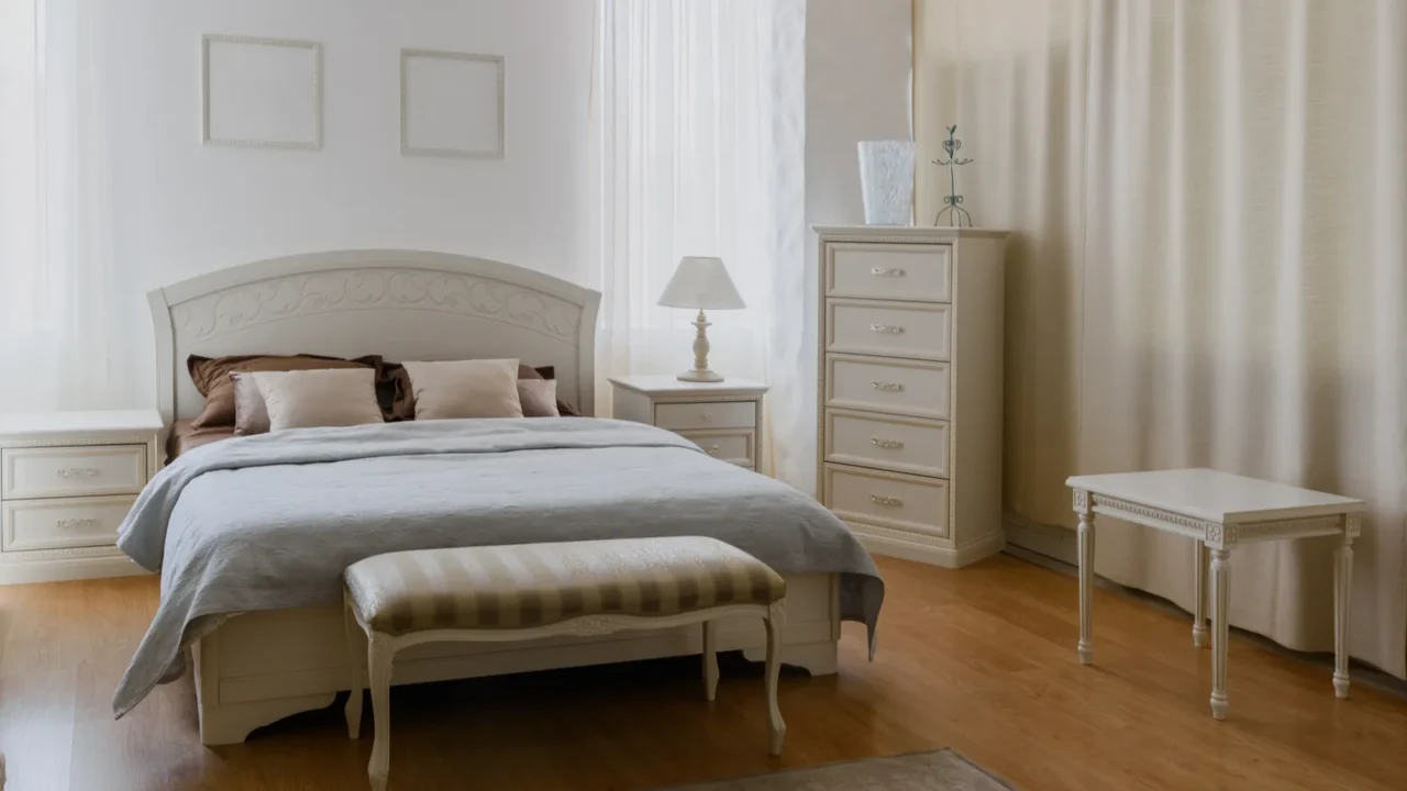 A beautifully styled bedroom featuring a white ornate bed with brown and beige bedding, elegant nightstands, a tall dresser, and soft natural light filtering through sheer curtains.