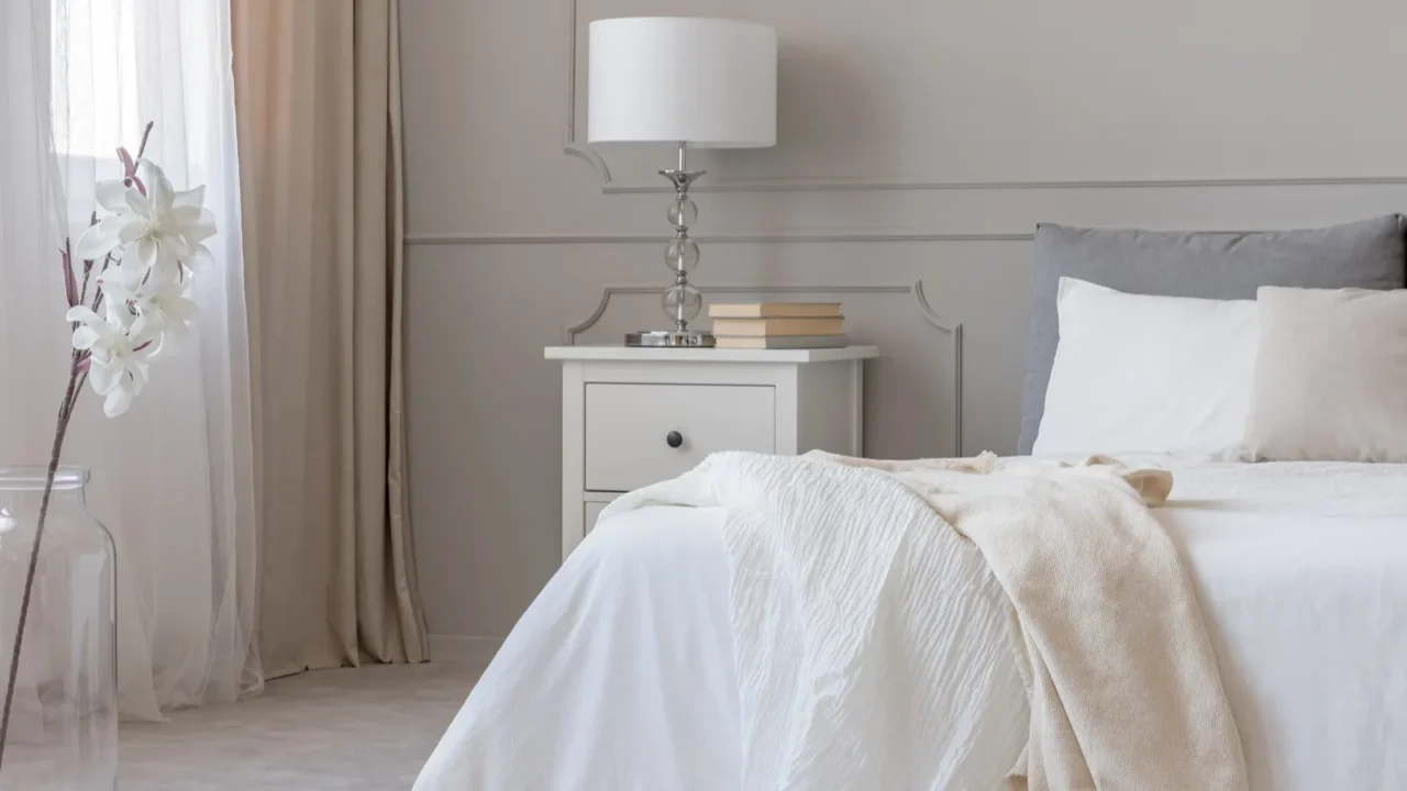 A bedroom interior with cozy bed and white nightstand with lamp and stack of books on it.