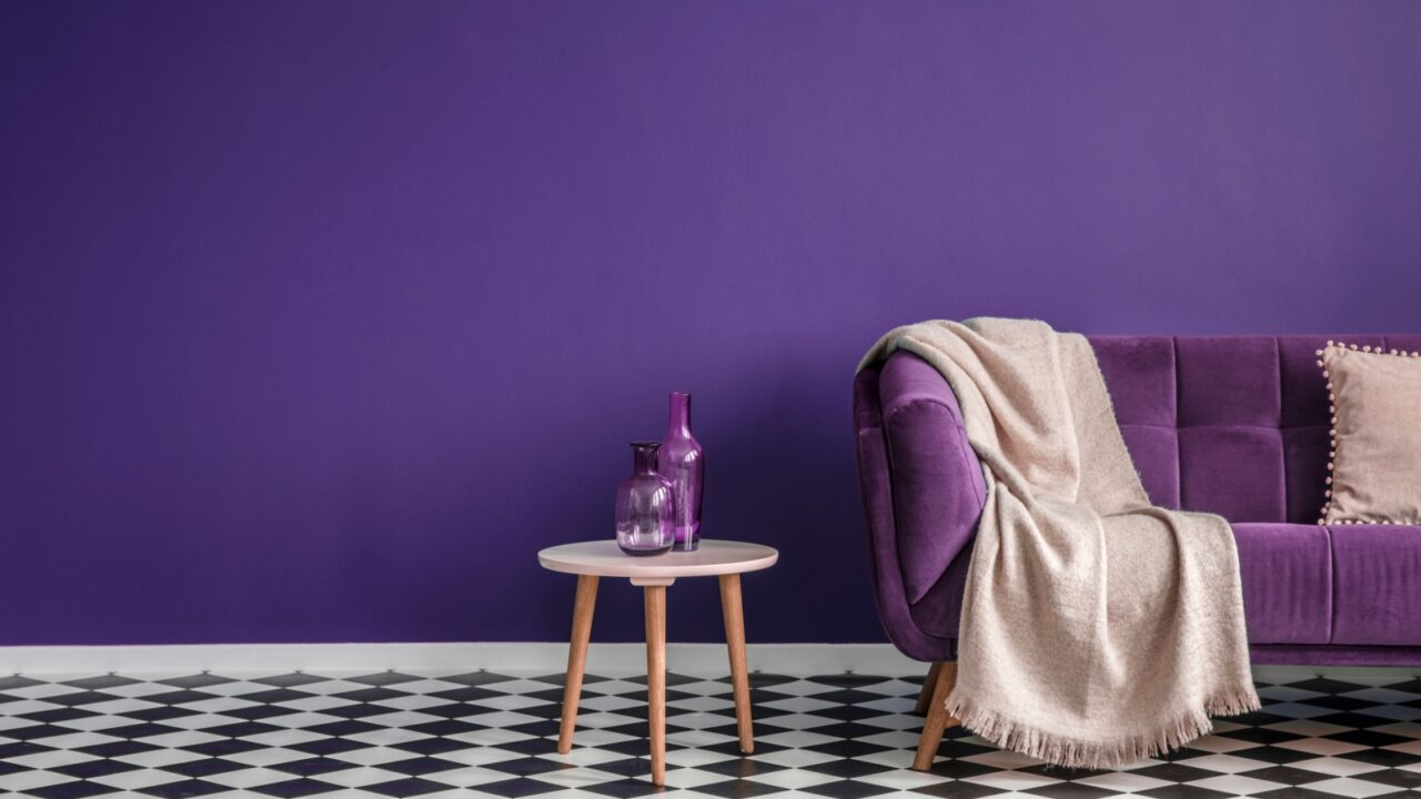 Dark purple sofa with a blanket beside a small table with bottles standing on black and white checkerboard floor in a minimalistic living room interior.