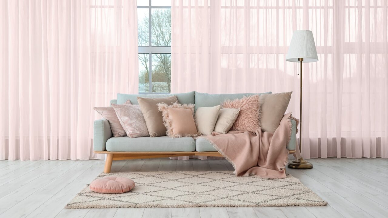 A living room interior with a cozy sofa with throw blanket and pillows, floor lamp, blush pink sheer curtains on the windows, and a rug.