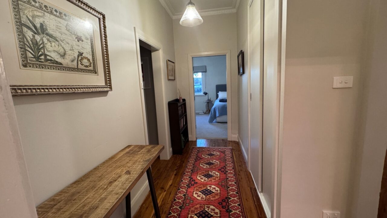 A hallway with a wooden floor, a red runner rug, a wooden console table, framed artwork on the walls, and a view into a bedroom.
