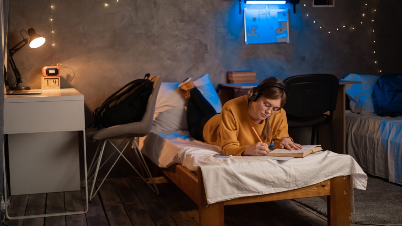 Room with warm lighting where a girl studies on a bed with books and notebooks scattered around.