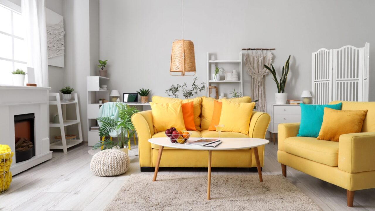 Interior of living room with yellow sofa, fireplace, woven light fixture, coffee table with fruits in a wire basket, and plants