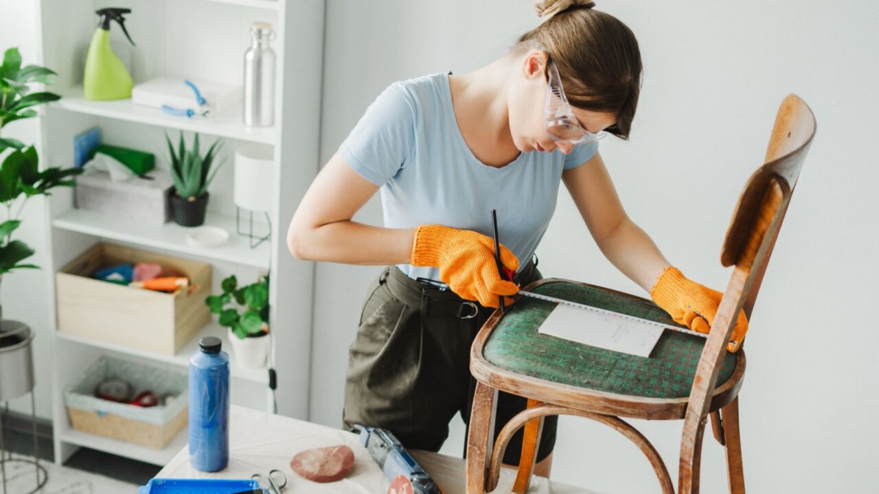 A woman taking measurements of a chair's seat using an inch tape. She is wearing casual outfit with protective glasses and orange gloves.