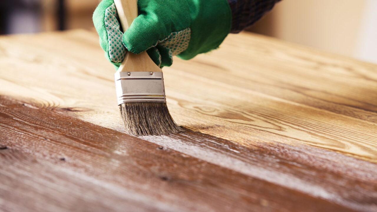 A person wearing green gloves applying dark wood stain on a wooden table using brush.