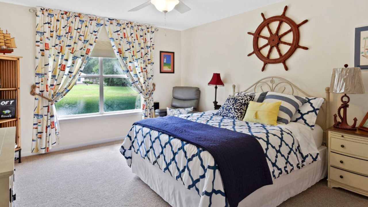 A bedroom interior with a cozy bed with patterned pillows and blanket, nightstand, lamp, patterned curtains, and a ship's wheel on the wall.