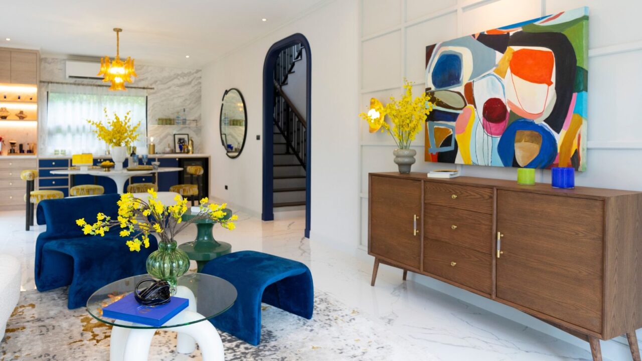 A living room interior with a wooden console, blue sofa, coffee table with flower vase, and a colorful oversized abstract art on the wall.