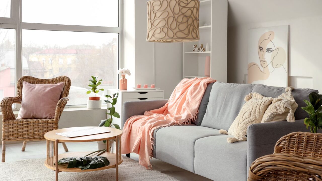 A stylish living room interior with a gray sofa with cozy throw pillows and blanket, wicker chairs, wooden coffee table on a rug, and wall art.