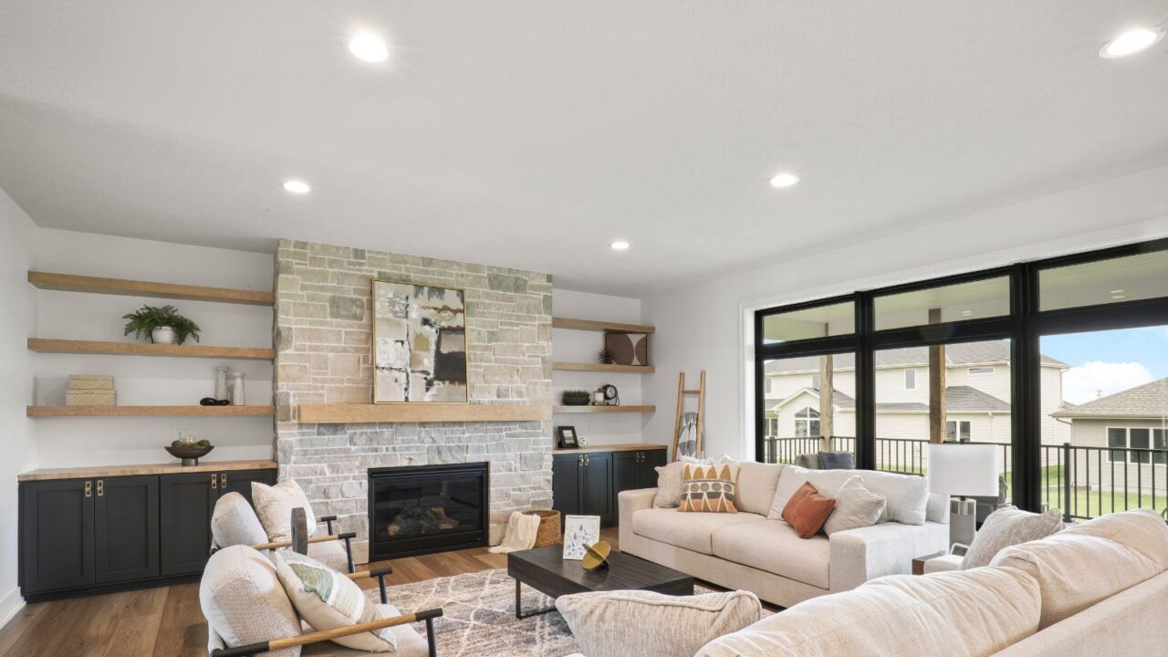 A stylish living room interior with cozy sofas, wooden floating shelves, fire place, and overhead ceiling lights.