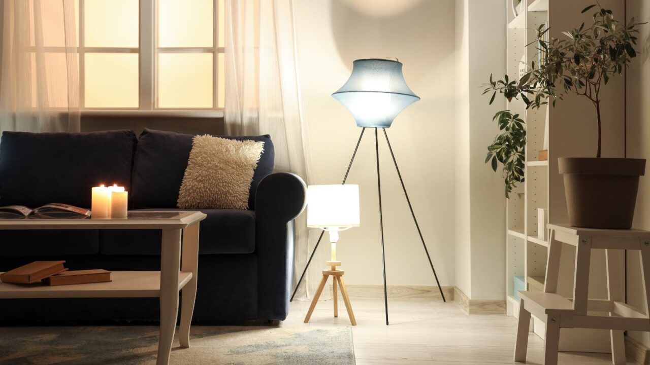 A living room interior with a blue sofa, throw pillow, coffee table with burning candles and books, houseplant, and floor lamps of different lengths.