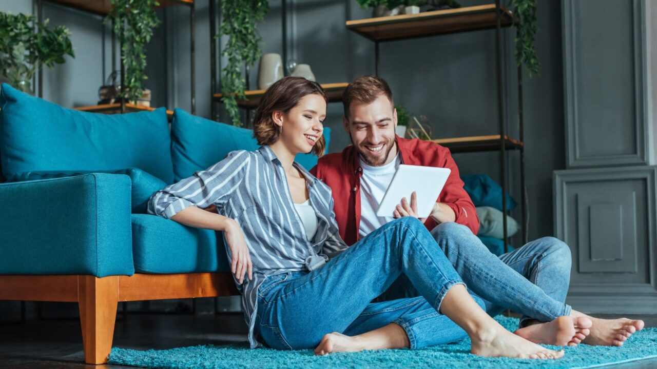 A happy couple is sitting on a rug in a living room. They are smiling while looking at the tablet in woman's hand.