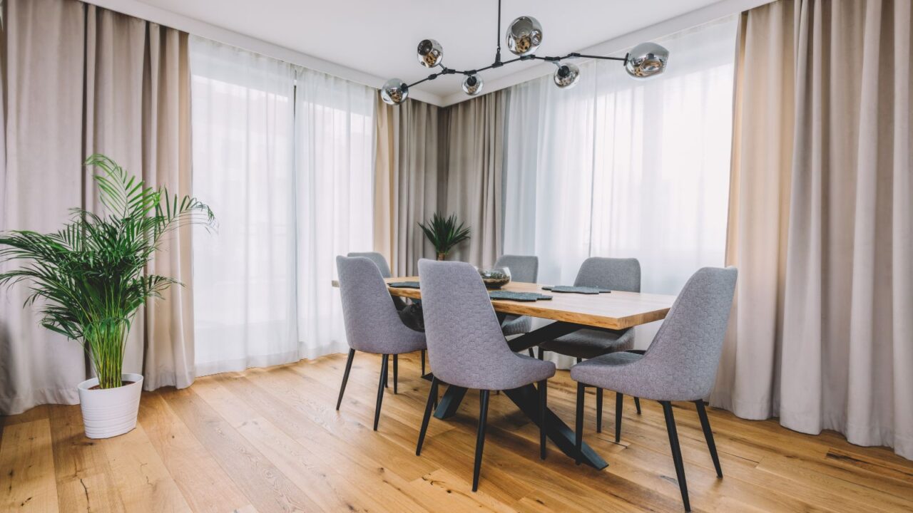 A stylish dining room interior with a wooden table, chairs, plant, layered curtains, and chandelier hanging from the ceiling.