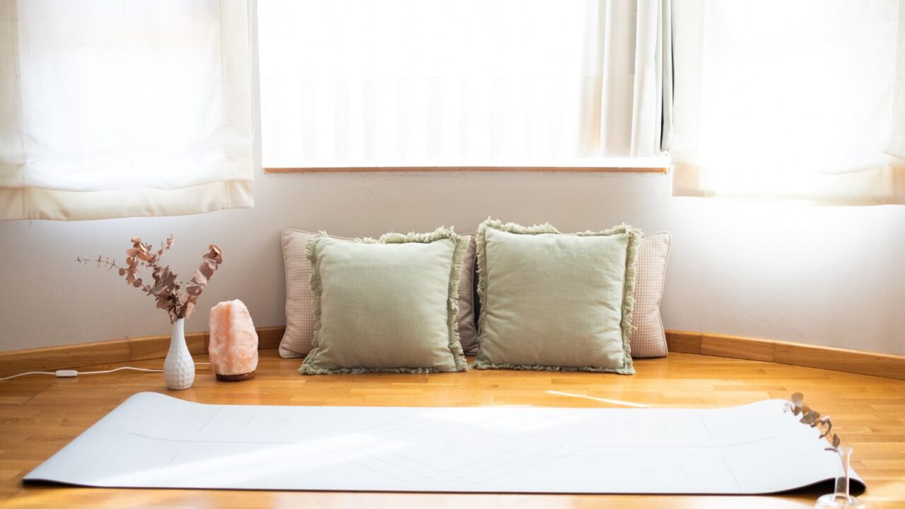 A meditation nook with a yoga mat, throw pillows, vase with dried leaves, and a Himalayan salt lamp on a wooden floor with sunlight filtering through the blinds.