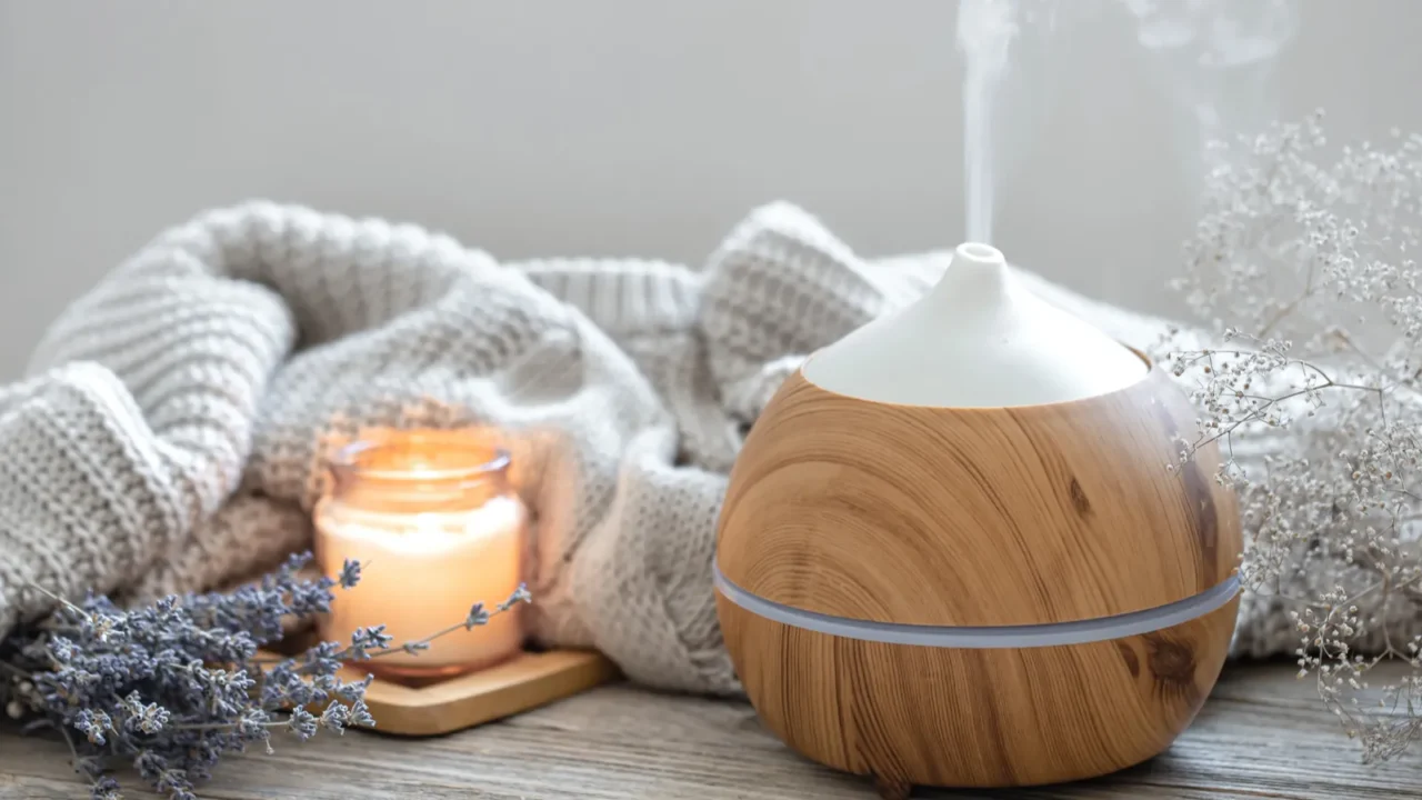 A modern aroma oil diffuser, knitted throw blanket, lavender, and burning candle on a wooden surface.