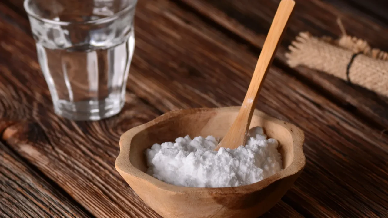 A glass of water and a wooden bowl filled with baking soda on a wooden table.