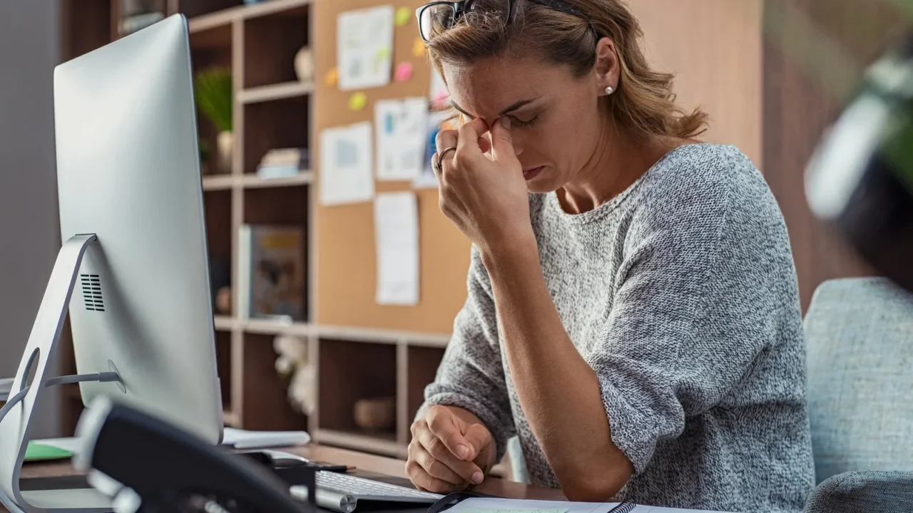 A woman sitting at a desk, stressed while rubbing her forehead in front of a computer monitor, with office supplies scattered around.