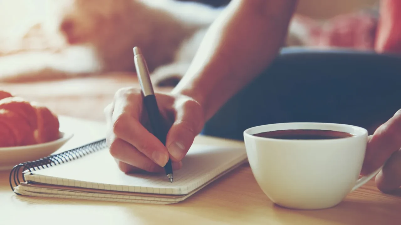 A woman writing on a dairy while holding a cup of coffee.