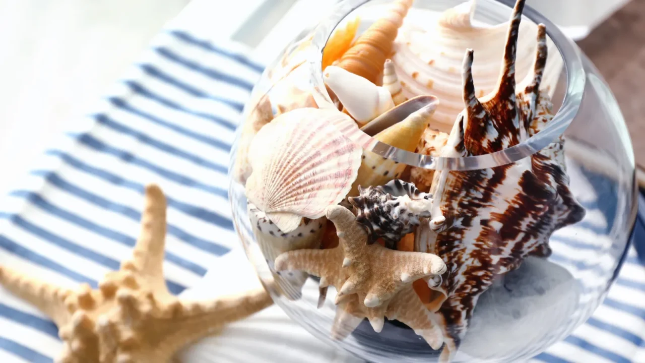 A glass bowl filled with various seashells and starfish placed on a striped cloth.