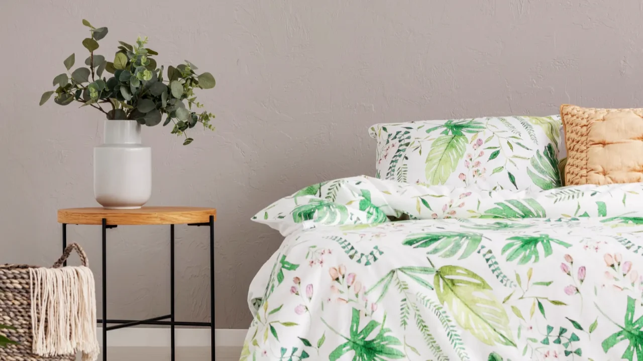 A bed with floral bedding with a wooden bedside table with flowers in a gray vase on it.