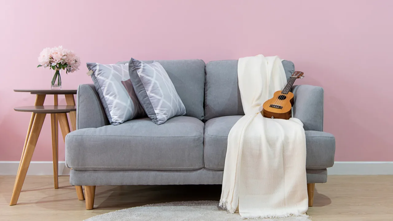 A stylish living room with a wooden gray sofa with pillows, white throw blanket, and a guitar, rug, and wooden coffee tables with a flower vase in front of a pink accent wall.