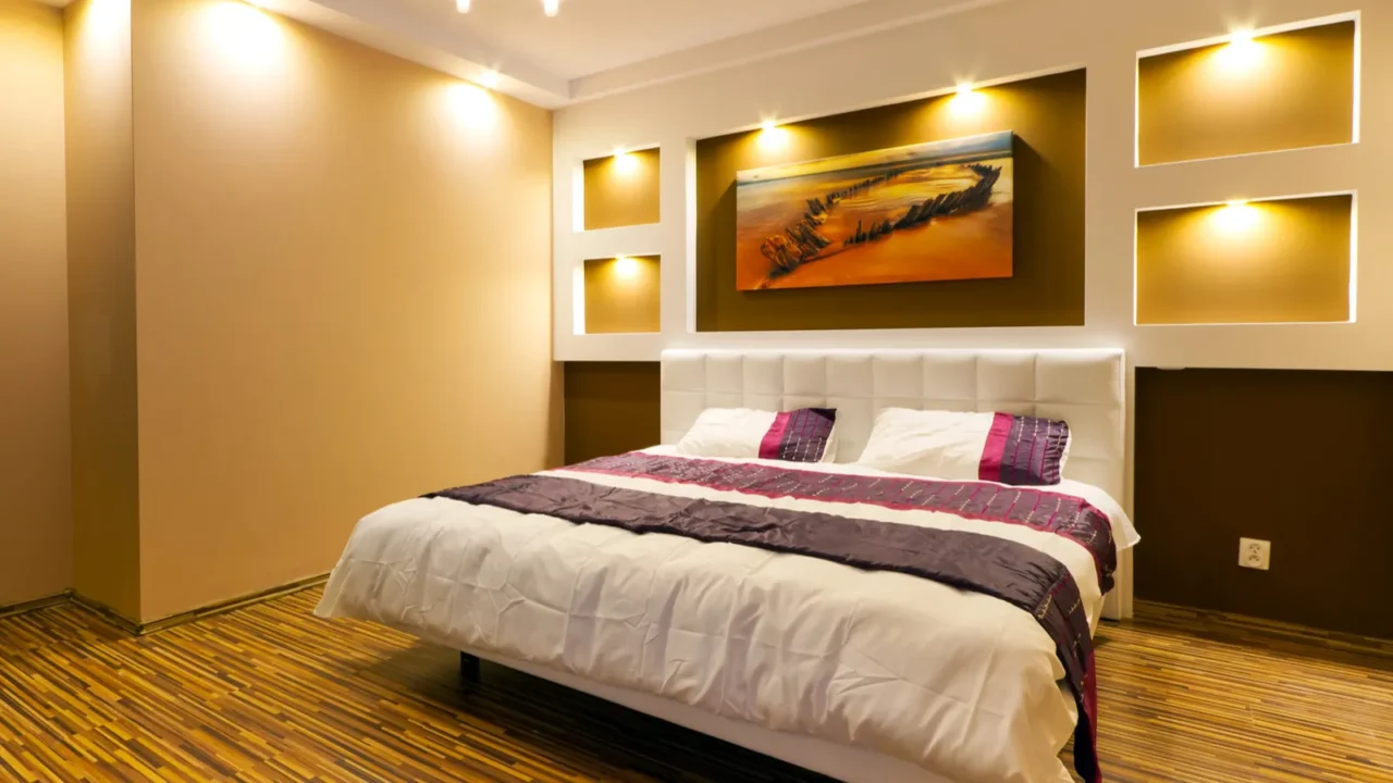 A bedroom interior with a cozy bed on a wooden floor, a painting on the wall, and warm toned light bulbs illuminating the room.