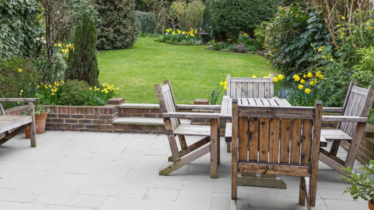 A wooden garden furniture set on a stone patio with blooming flowers.