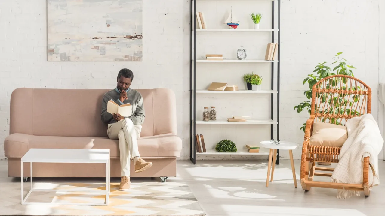 A stylish living room with a man reading a book while sitting on a cozy sofa, a shelf with books and decor accents, rattan armchair with throw pillow and blanket, and plants.