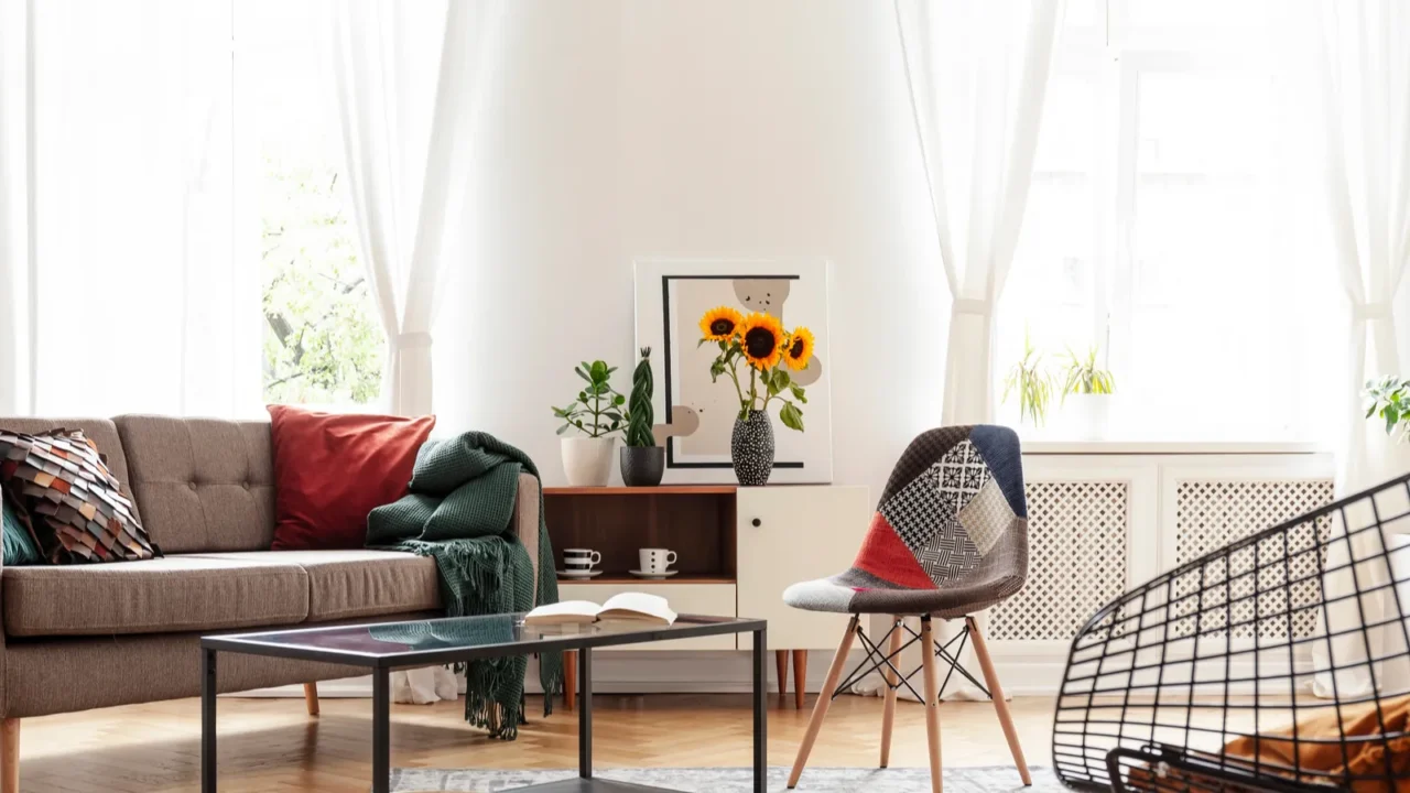A white living room interior with brown sofa with colorful cushions and throw, coffee table, accent chair, and white sheer curtains on the windows.