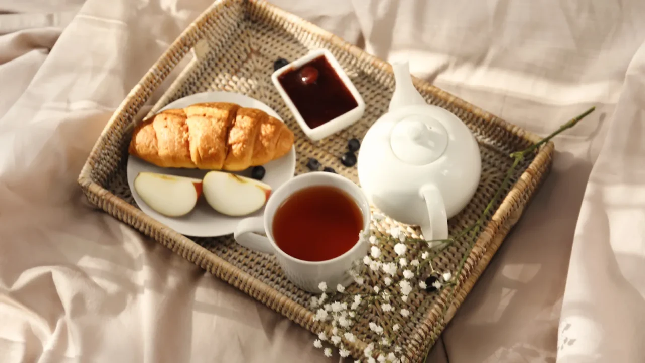 A breakfast tray with croissant, boiled eggs, tea, and jam on a bed.