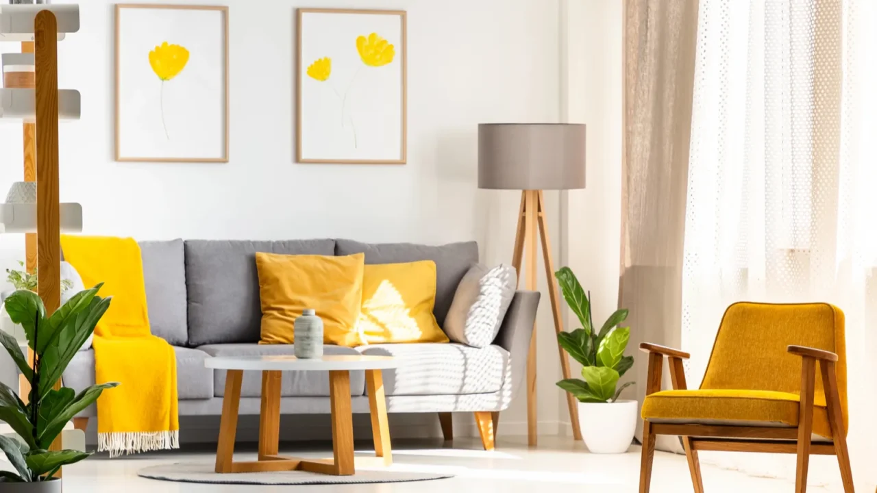 A stylish living room interior with a cozy gray sofa with yellow throw blanket and pillows, a wooden yellow armchair, floor lamp, plants, and framed artwork on the wall.