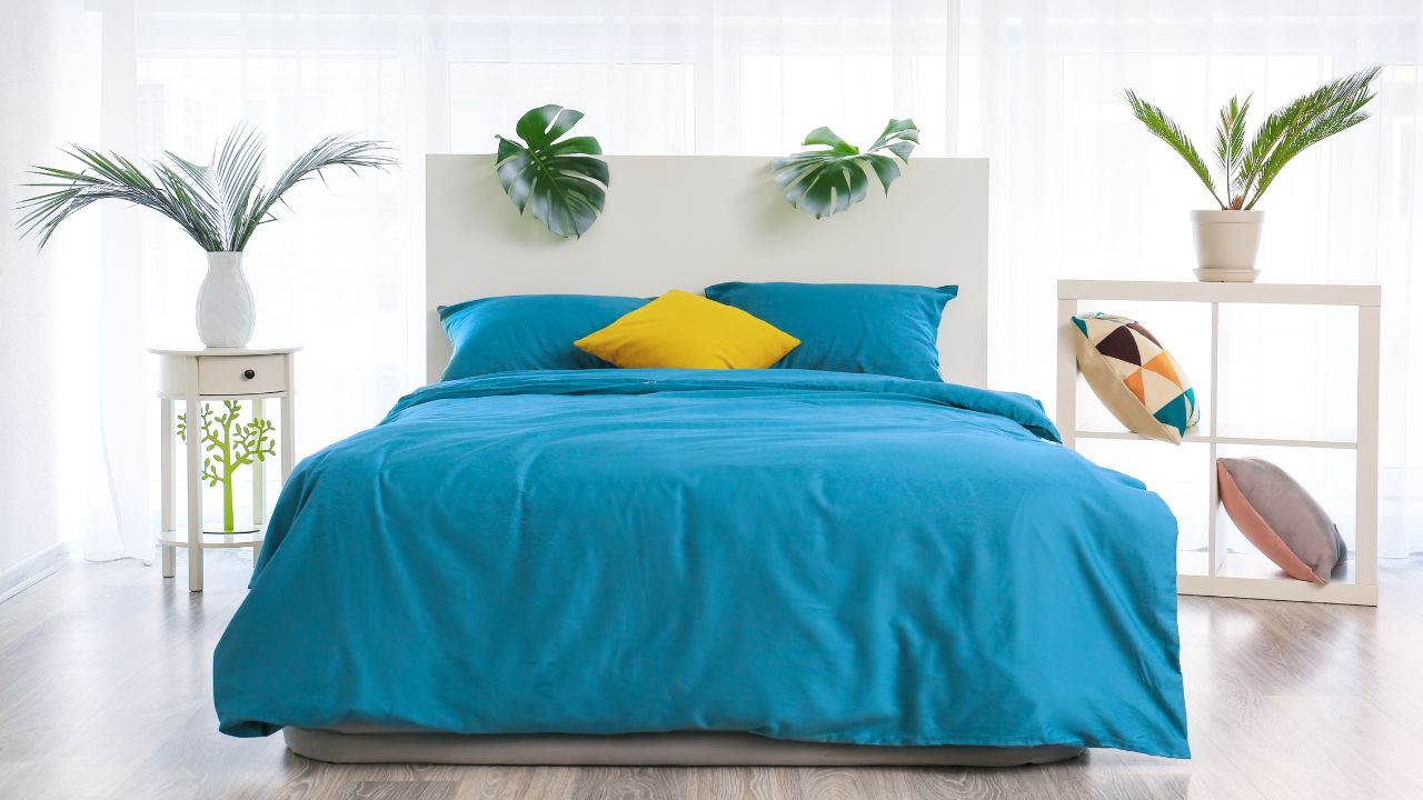 A bedroom with a blue bedspread, yellow pillow, white nightstands, and potted plants.
