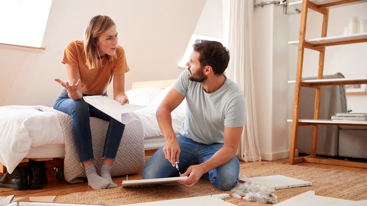A woman and a man are having a conversation on the floor of a room under renovation, surrounded by tools and assembly parts.