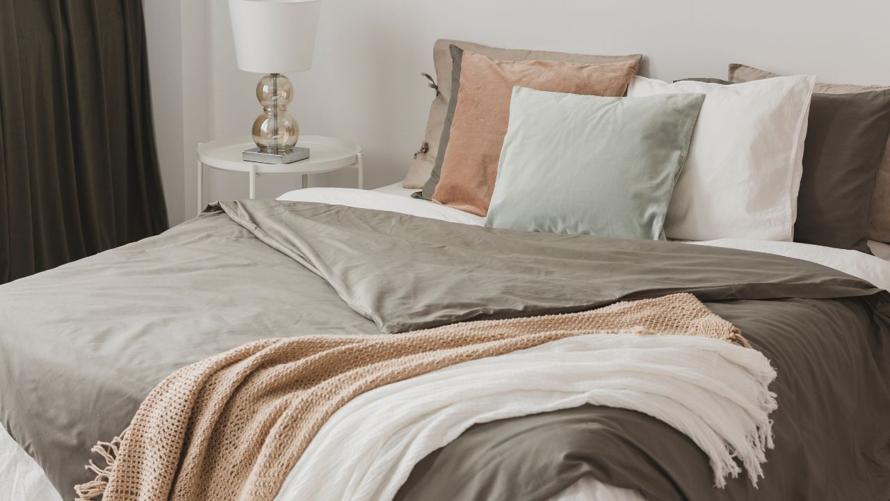 A neatly made bed with gray and white bedding, multiple pillows, and a bedside table with a lamp.