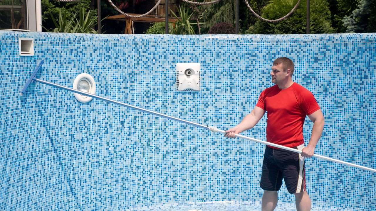 Person cleaning a blue mosaic-tiled pool with a long pole.