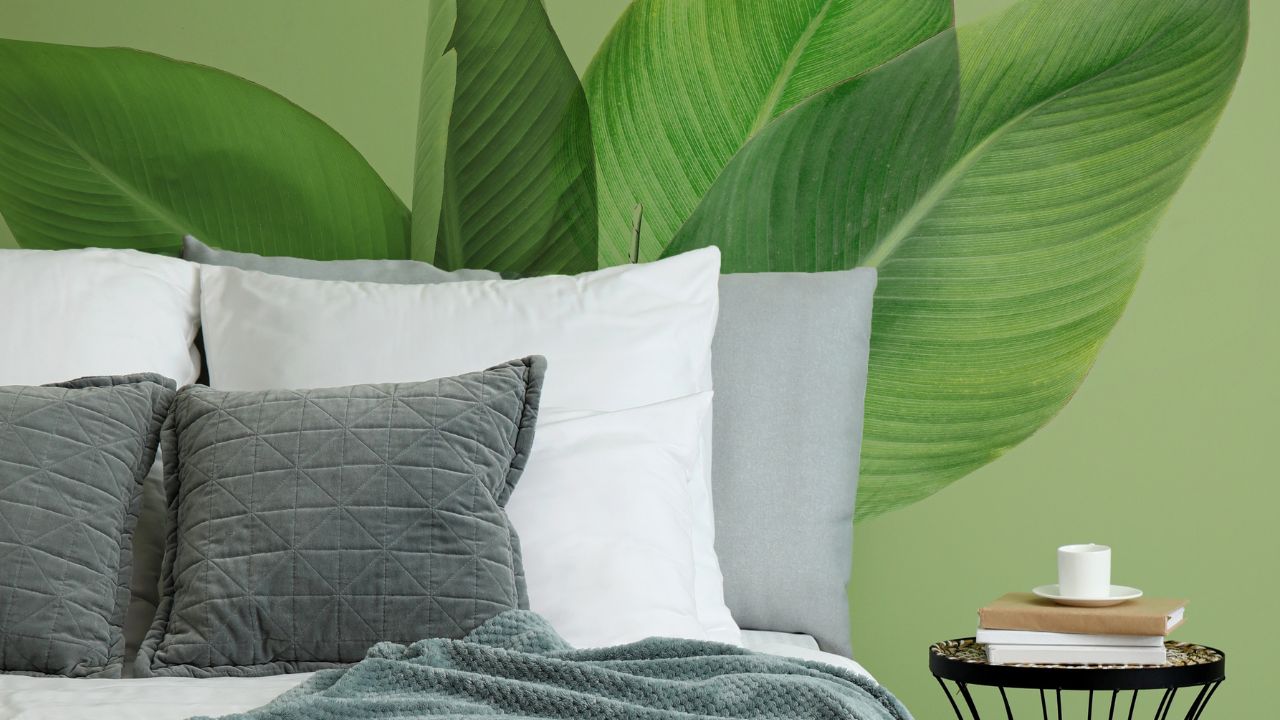 A cozy bed with white pillows, gray cushions, and a textured blanket, with a large green leaf mural in the background.
