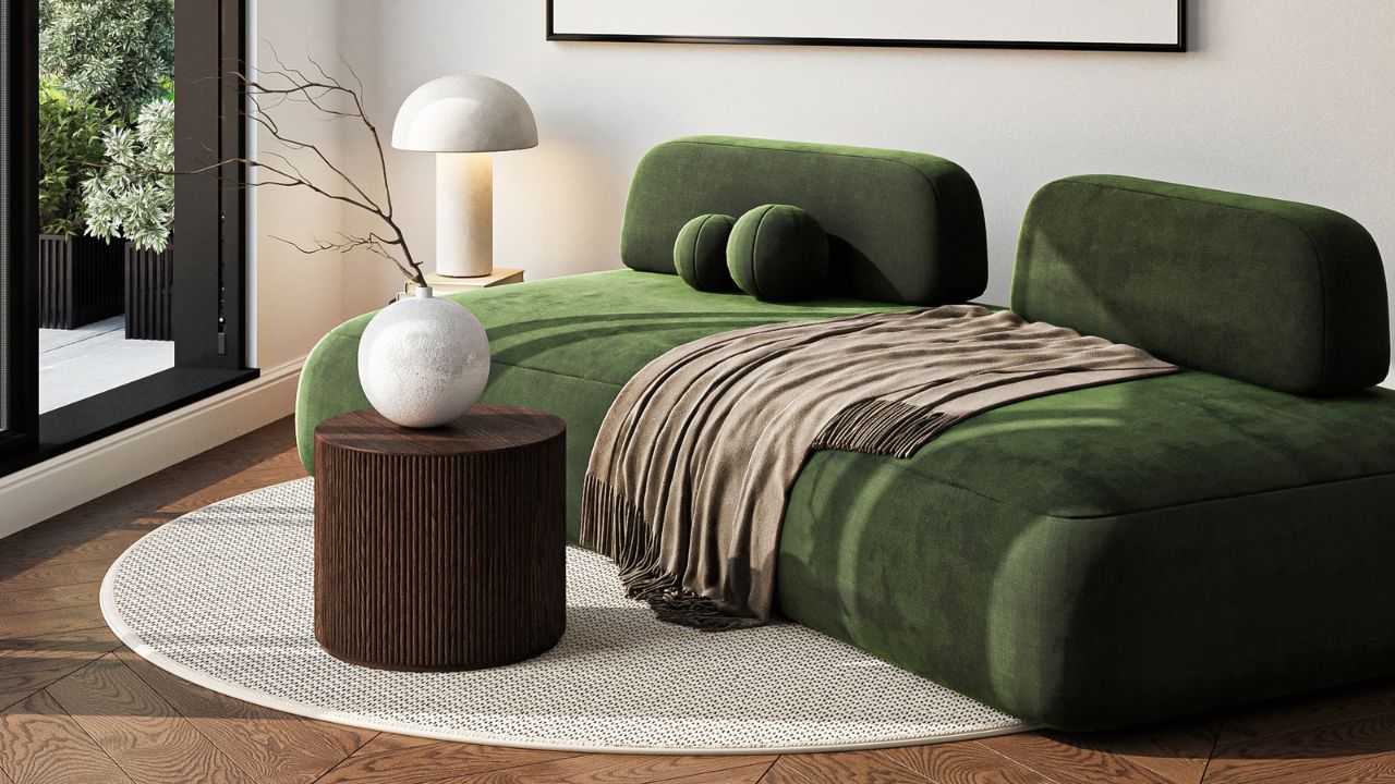A modern living room corner with a green upholstered sofa, a round wooden side table with a white vase, and a white lamp, next to a large window with greenery outside.