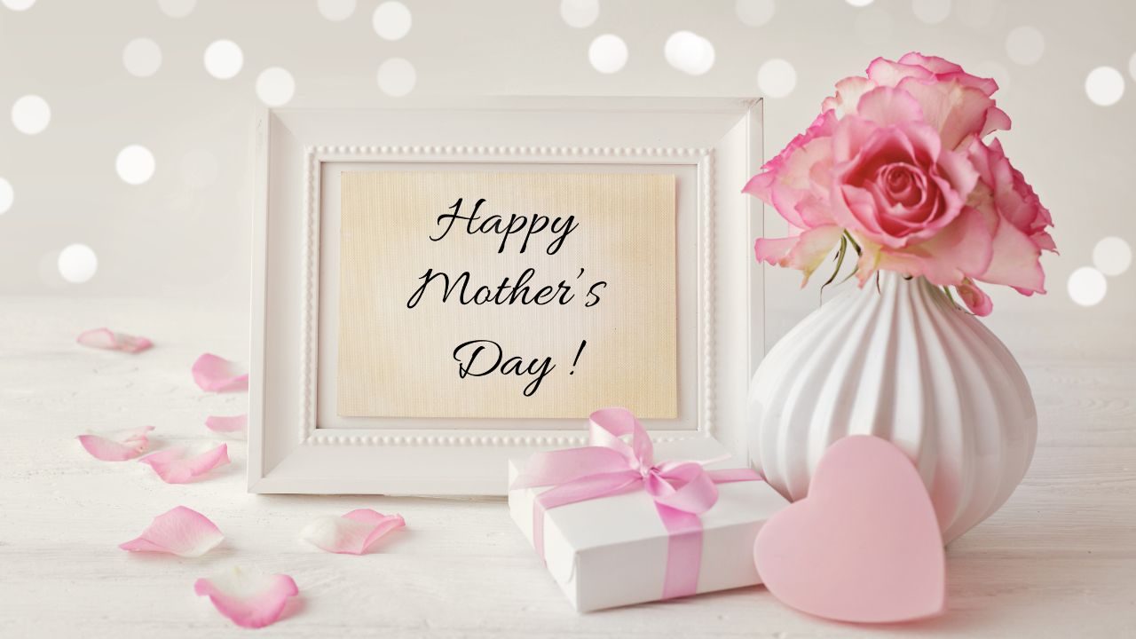 A Mother's Day greeting setup with pink roses, a gift, a heart, and a framed message on a white surface.