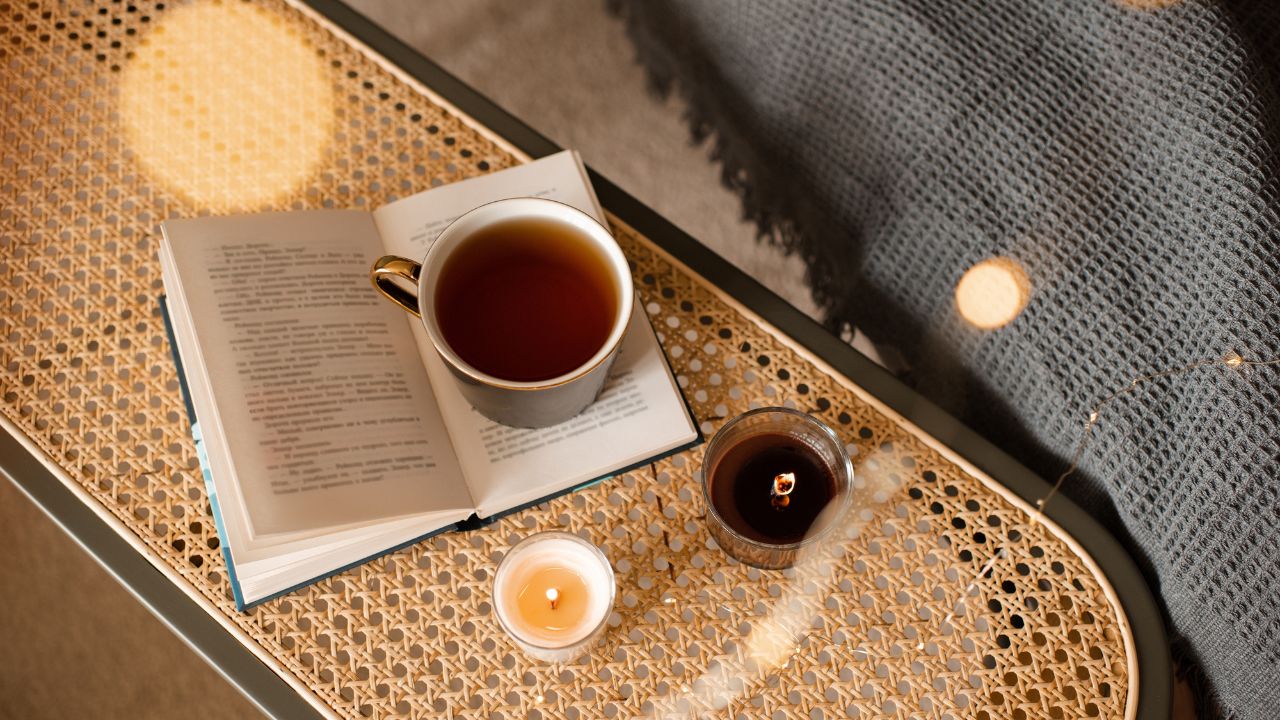 A close-up of a table with an open book, a cup of tea, and a lit candle.